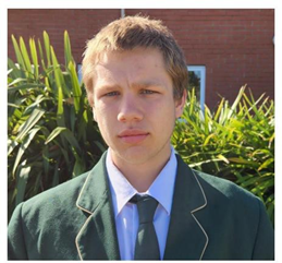 Aiden Mill, wearing a green blazer, stands in front of a flax bush