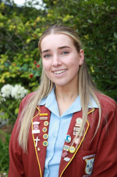 Maevi Fleming smiles in front of an array of shubbery and flower bushes. She wears a red school blazer with various pins and badges