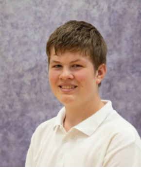 Sam Nicholls sits side on, smiling in front of a plain background