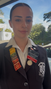 Sophie stands in front of a large window, smiling and wearing her school blazer covered in badges and pins