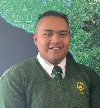 Taiko smiles at the camera in front of a map of New Zealand wearing a green school uniform sweatshirt.